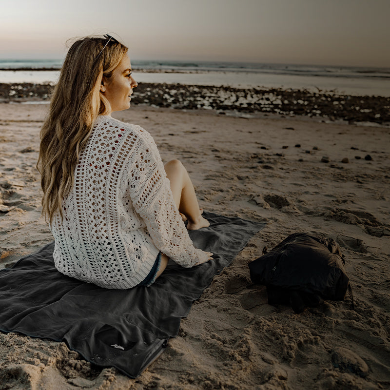 A woman sits on a beach blanket, wearing a white knitted sweater, with the ocean and sunset in the background. A backpack is nearby.